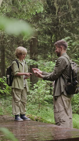 Vertical shot of 9-year-old Caucasian boy spraying insect repellent on dads hands while going wild camping together in summer nature