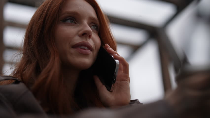 Low-angle view of positive young woman with fiery red hair talking on smartphone swinging on city swings, holding coffee cup, radiating joy and self-assurance in contemporary city environment