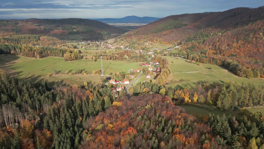 Mountains covered with colored autumn trees and small village in valley, aerial view. Beautiful nature landscape in fall season