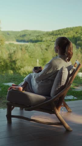 A calm and relaxed woman sitting in a comfortable chair on the terrace of a country house, drinking tea and enjoying view of nature. The concept of mental health and social distancing