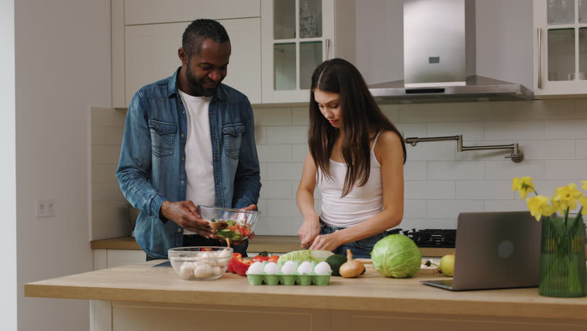 Couple is preparing meal together in modern kitchen. Woman is chopping vegetables while man is mixing salad in bowl. Countertop is filled with fresh ingredients. They following recipe online on laptop