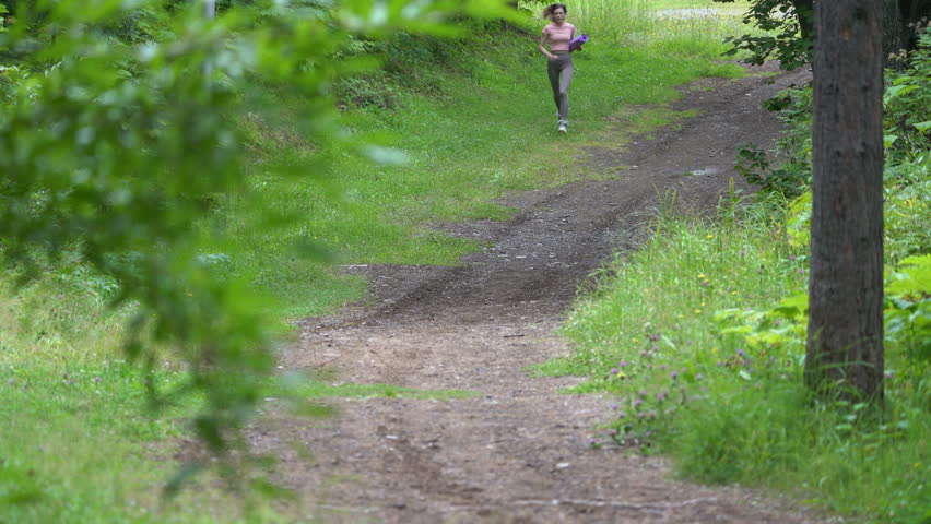 Front view of slender woman athlete jogging along footpath in park during physical activity, summer workout. Slim runner wearing sportswear and carries exercise mat. Full length