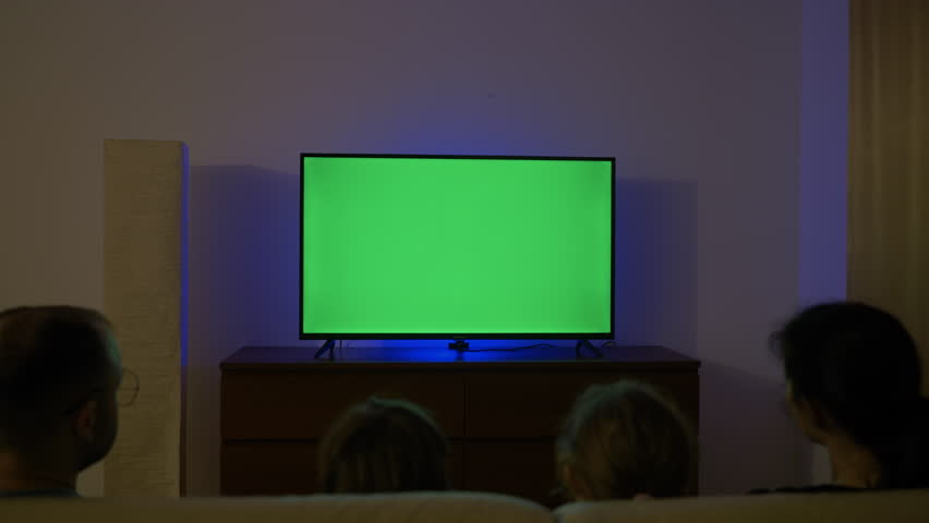 Family With Children Sitting Together Sofa In Their Living Room Night Watching TV Green Screen. Rear View Of Family Sitting On Sofa In Living Room Evening Watching Green Mock-up Screen TV Together.