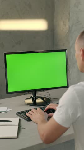 Young Man Working In Office On Desktop Computer with Green Screen Mock Up Display. Over the Shoulder- Close-up of Monitor With Green Screen Chromakey