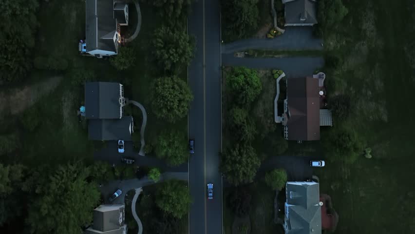 Aerial top down of car on straight road of upscale neighborhood houses with swimming pool. Large Real Estate homes in suburbia of USA. Dusk scene with green trees in summer. Birds eye tracking shot.