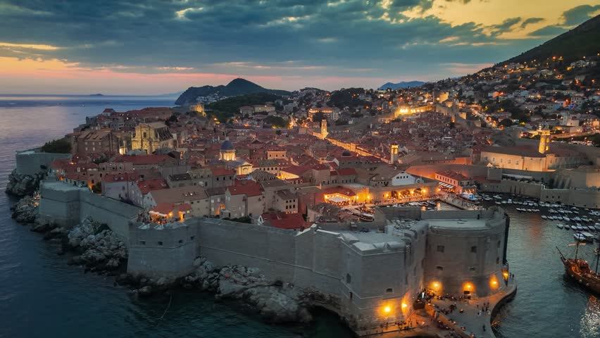 Dubrovnik old town after sunset with city lights. Aerial shot of historic city of Dubrovnik in Croatia, UNESCO World Heritage site. Illuminated streets, buildings and marina with boats. 4K HDR shot 