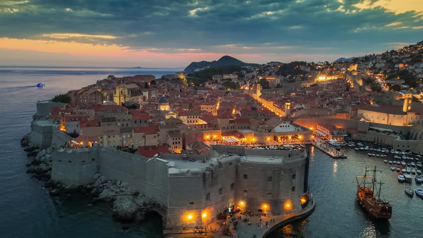 Dubrovnik old town after sunset with city lights. Aerial shot of historic city of Dubrovnik in Croatia, UNESCO World Heritage site. Illuminated streets, buildings and marina with boats. 4K HDR shot 