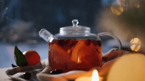 A glass teapot with whirling warm tea, cranberries, and citruses on a cozy table with a flickering candle nearby. Winter holidays, snow falling outdoors - Powered by Shutterstock - Get 15% off with code: PIKWIZARD15