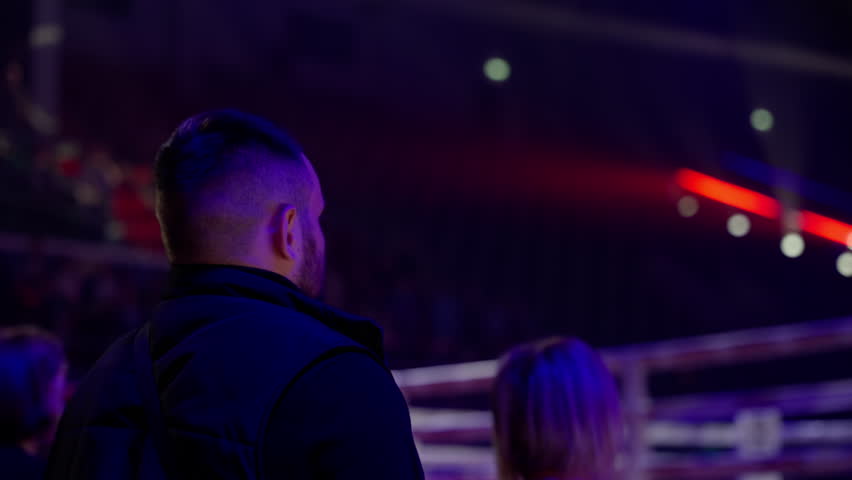 Security guard monitoring a sporting event in an arena, surrounded by dim and stage lighting. The atmosphere buzzes with excitement as the crowd anticipates the competition