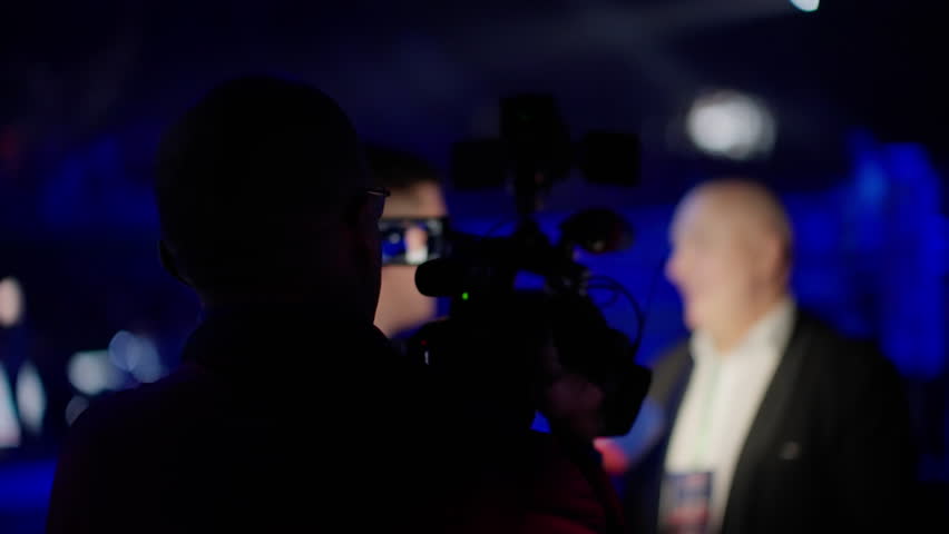 Cameraman capturing an interview with a businessman at a sporting event in an indoor stadium. Dim lighting enhances the dramatic atmosphere, emphasizing communication and media coverage