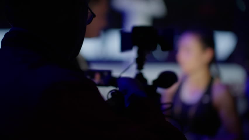 Videographer is filming a female athlete at a sporting event. The athlete is out of focus and the videographer is a silhouette