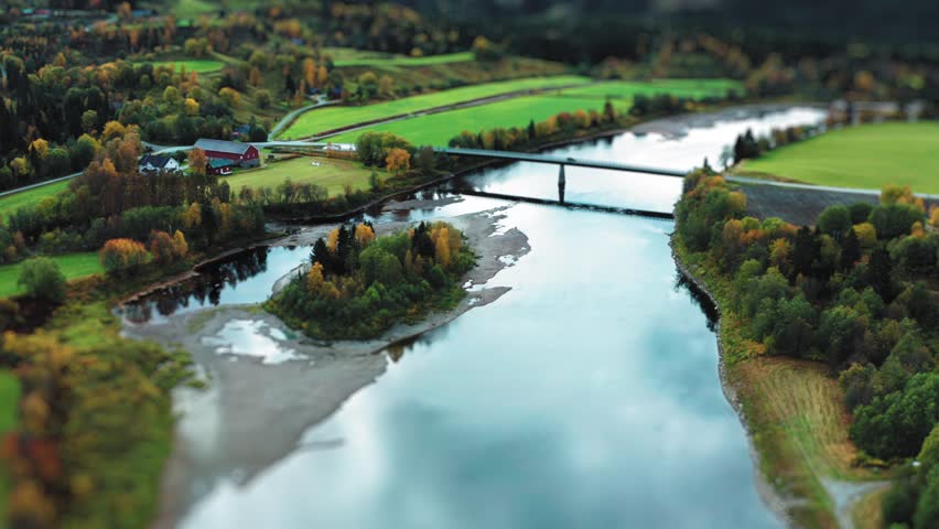 A bridge crosses over a winding river, surrounded by fields and trees dressed in early autumn colors. Aerial view.