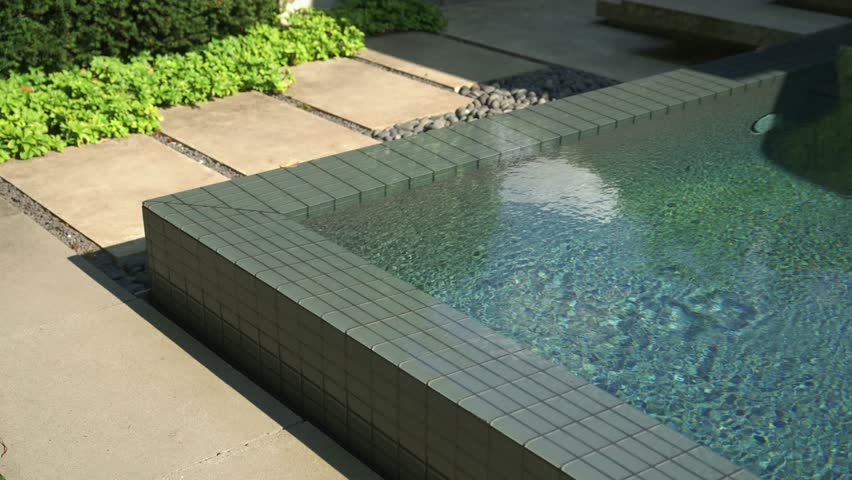 Slow orbit around the edge of a private luxury swimming pool in rear garden on bright sunny day with blue water rippling in the wind on the surface.