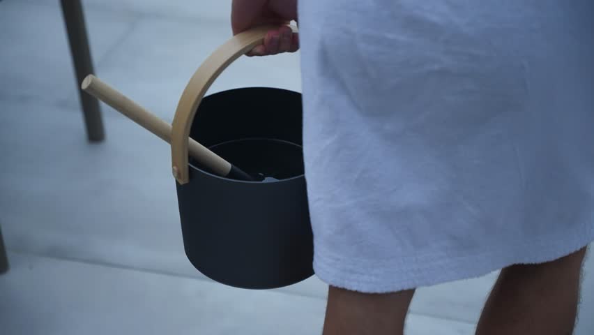 Close up of a white man dressed in a white dressing robe carrying a premium metal sauna water bucket with a wooden spoon walking down stairs at dusk