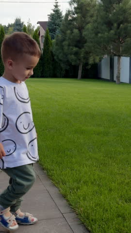 Cute Caucasian boy goes on the grass and steps by it turning around. Lovely baby spending time outdoor in summer. Vertical video.