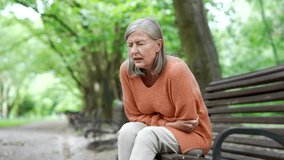 Elderly female with gray hair suffering from stomach pain sitting on bench on street in urban city park. Upset mature woman has problems with digestion, poisoning, gastritis, stomach ulcer or bloating - Powered by Shutterstock - Get 15% off with code: PIKWIZARD15