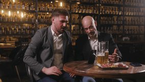 Two men, dressed in business casual attire, are sitting at a bar table, engaging in an intense conversation while looking at a smartphone propped up on the table. - Powered by Shutterstock - Get 15% off with code: PIKWIZARD15