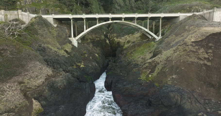 Aerial pullback from concrete bridge at Captain Cook Point, Cape Cove, Oregon Coast as waves crash and spray mist up into air