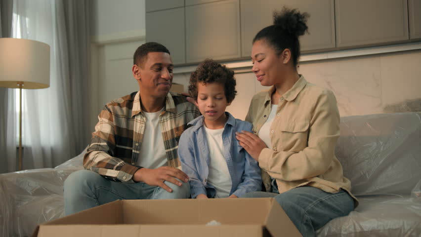 Little kid boy adopted child open cardboard box parcel surprise gift birthday present toy excited happy son hugging teddy bear parents children African American bonding love family at home moving day