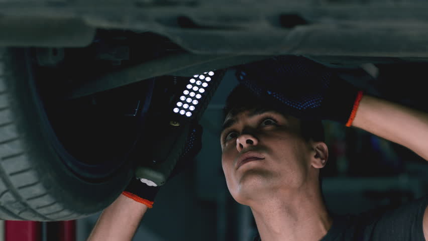 Car repair service. Young auto mechanic holding light and examining vehicle suspension in garage, close up