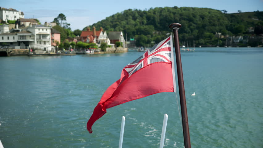 Great Britain Red Ensign flag fluttering in the wind on stern of ship with sea or lake and a small town in the background.