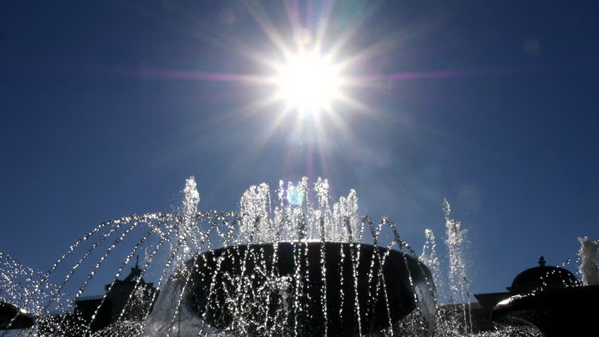 Pigeons fly over a fountain against the backdrop of a sparkling sun in a clear blue sky on a hot summer day.