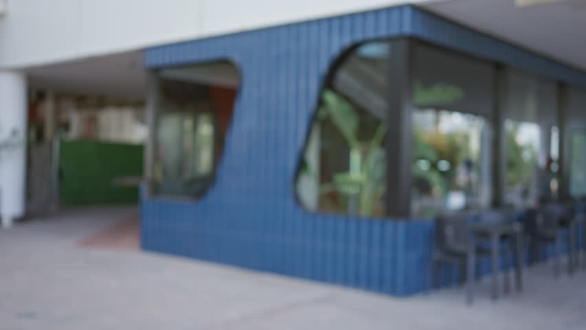Blurred outdoor cafe with blue wall and glass windows, featuring defocused plants and chairs in the background.