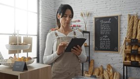 Hispanic woman in a bakery, wearing an apron, using a tablet, surrounded by pastries, bread, and a chalkboard menu. - Powered by Shutterstock - Get 15% off with code: PIKWIZARD15
