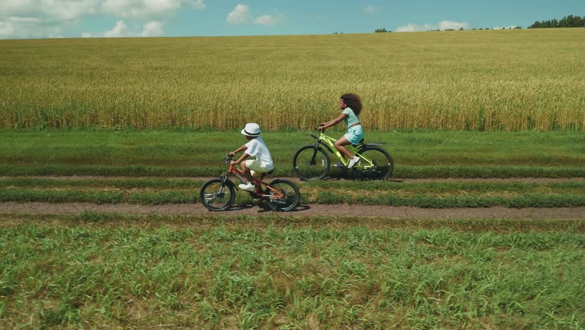Two children joyfully ride their bikes along a dirt path, surrounded by lush greenery and a golden field under a bright blue sky. It