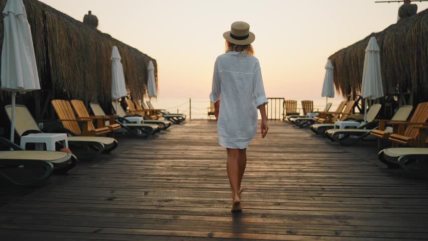 Barefoot woman walking in sunrise on sea pier, turning to camera, back view. Happy lady admiring sunset above sea horizon, carefree beautiful female person in nature, merry summer trip, slow motion
