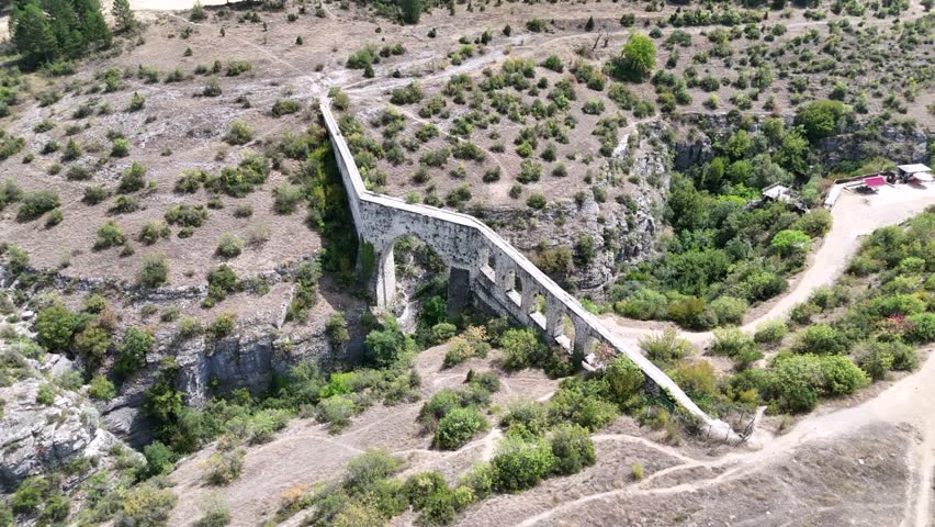 Against a cloudy but clear sky, a mountain view features a river and canyon. In the center, a historic Roman bridge or arch stands out, blending ancient architecture with stunning natural scenery