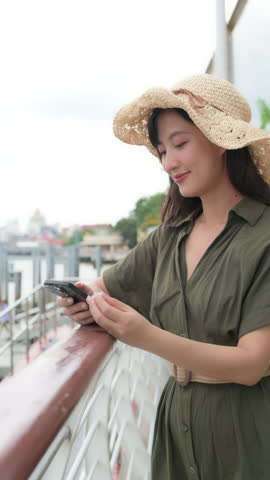A young woman enjoying music on her phone, smiling with relaxed expression, wearing straw hat and green dress. background features scenic waterfront. Vertical video.