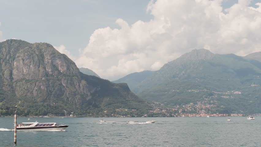 A boat crosses the waters beneath the steep, imposing mountains of The Alps in Lake Como, Italy.