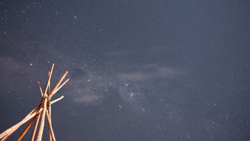Timelapse made of the rotating Milky Way moving above some wooden pointy tipi sculpture in the foreground at night in Chili South America with falling stars across the video and small clouds pasing by