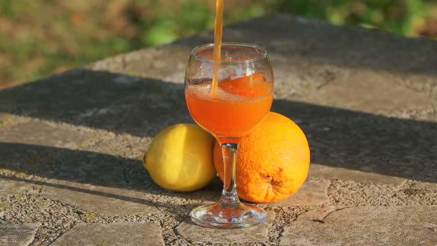 Slow motion of pouring orange juice into glass. Placed on stone table with green garden in the background.
