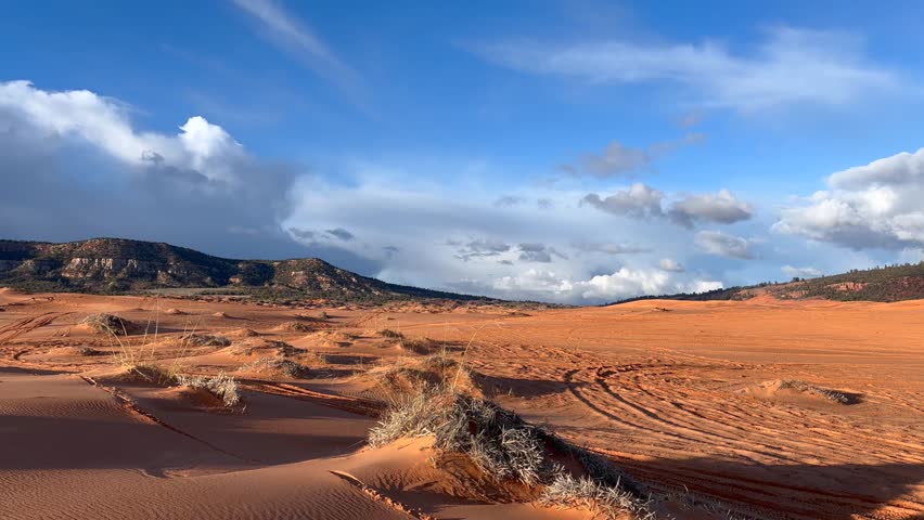 An ATV drives through a valley of sand dunes with at sunset mountain range and clouds in the background at Coral Pink Sand Dunes State Park in Utah, USA.