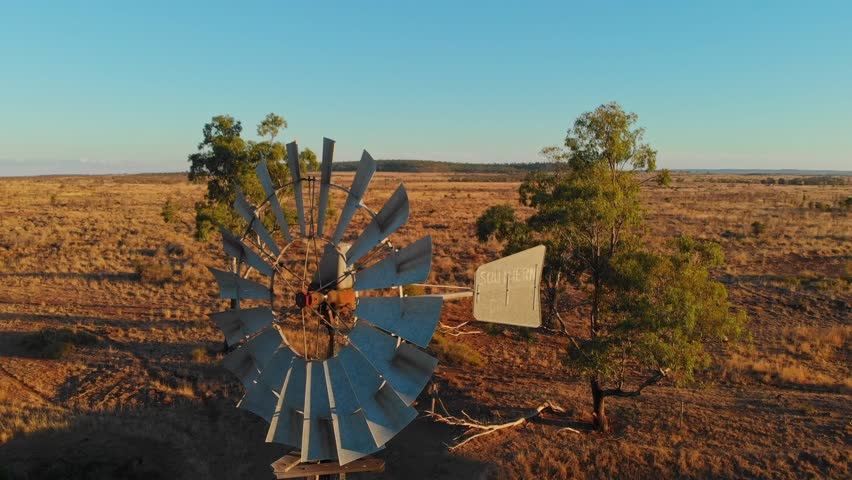Aerial view of a serene sunset over a vast dry landscape with an old windmill and trees, Emerald, Queensland, Australia.
