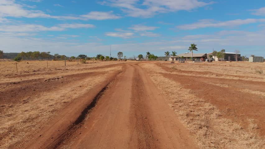 Aerial view of a tranquil rural landscape with a farmhouse, barn, and palm trees amidst dry fields and dirt roads, Emerald, Queensland, Australia.