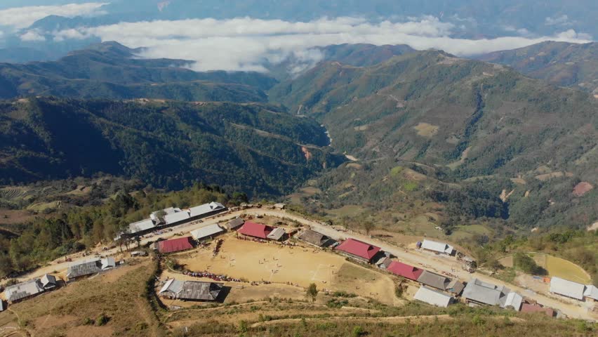 Aerial view of a serene village with a soccer field and houses amidst mountains and clouds, Sin Ho, Vietnam.