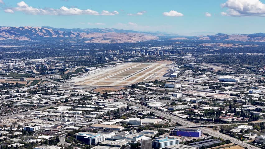 Aerial view of San Jose International Airport with surrounding urban landscape and distant mountain ranges under a clear sky in Silicon Valley, California.
