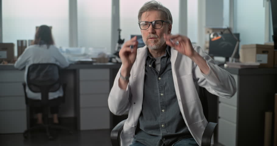Mature scientist in lab coat speaks to the camera and gives interview for popular science film sitting in archaeological scientific laboratory. Female colleague works in the background. Static Shot.