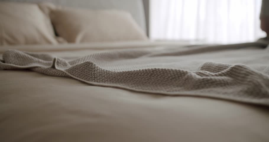 Close-up of a cozy bed featuring a textured blanket and neutral tone bedding, bathed in natural sunlight through sheer curtains, creating a serene and inviting atmosphere. Cozy Bed with Soft Blanket