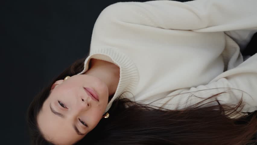Beautiful young brunette woman with white sweater and pants posing at studio. Beautiful woman having smooth healthy skin. Portrait of gorgeous woman looking to camera. Vertical video.