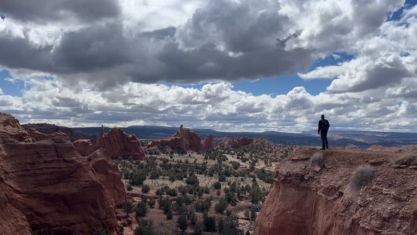 A man stands on a cliff edge in the desert, looking out at the amazing rock formations and vast landscape. The cloudscape is stunning against the blue sky. Kodachrome Basin State Park, Utah, USA