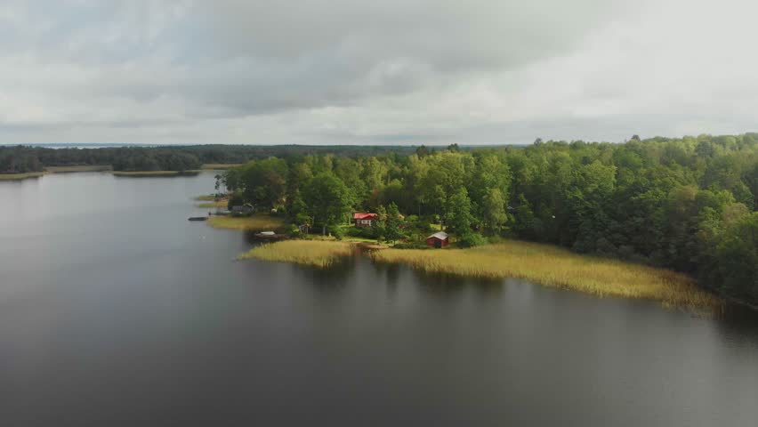 Aerial view of a serene lake surrounded by a tranquil forest with a picturesque traditional red wooden house, Ljungby, Sweden.