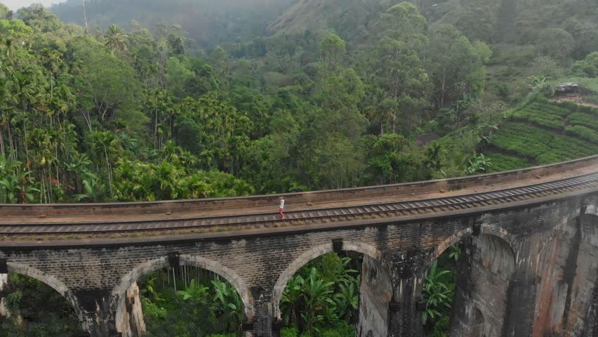 Aerial view of the nine arches bridge with a man dancing amidst lush tea fields and a cloudy sky, Ella, Sri Lanka.