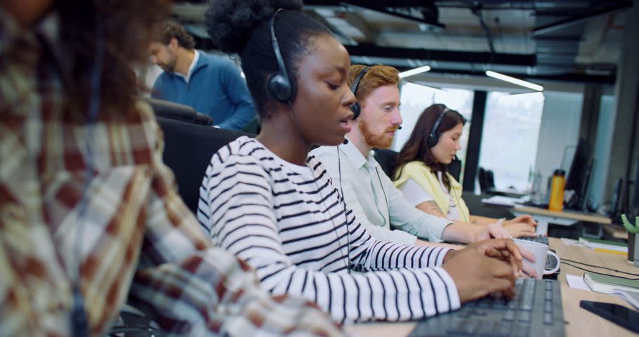 Professional multi-ethnic managers working in call center office. People sitting in row at desk and using computers. Discussing something with customers on headset. Team of workers answering calls.