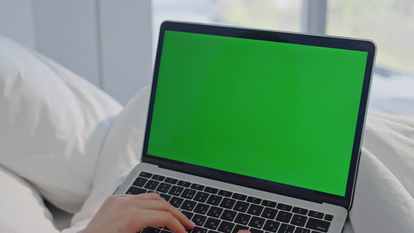 Closeup hands typing green screen laptop lying bed closeup. Unknown woman working on chroma key computer at calm morning home. Relaxed businesswoman browsing mockup pc in serene bedroom apartment.