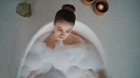 Peaceful lady relaxing in bubbles bath touching gentle skin top view. Calm woman lying in spa bathtub with white foam surrounded aroma candles. Beautiful girl in luxury bathroom practicing self care. - Powered by Shutterstock - Get 15% off with code: PIKWIZARD15