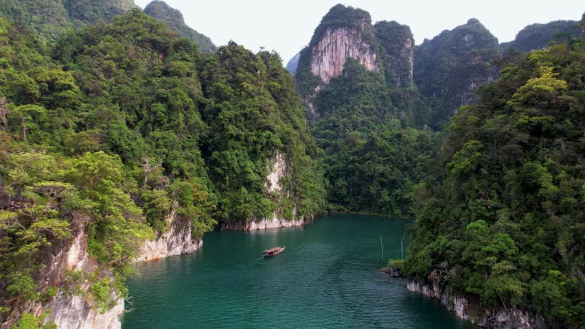 4K drone video flying towards a boat sailing around Khao Sok National Park in Thailand. This natural paradise os packed with towering cliffs and dense jungle. The boat is sailing all alone.